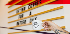 White cup of coffee with Ragamuffin Coffee Roasters logo in front of brand sign that says "Better Coffee for a Better Day."
