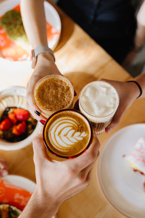 Three different coffee drinks coming together over a table of food at a coffee shop